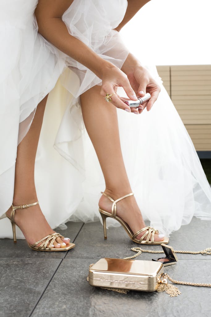 Crop unrecognizable bride in white dress and high heeled shoes holding lipstick in hands while preparing for wedding ceremony