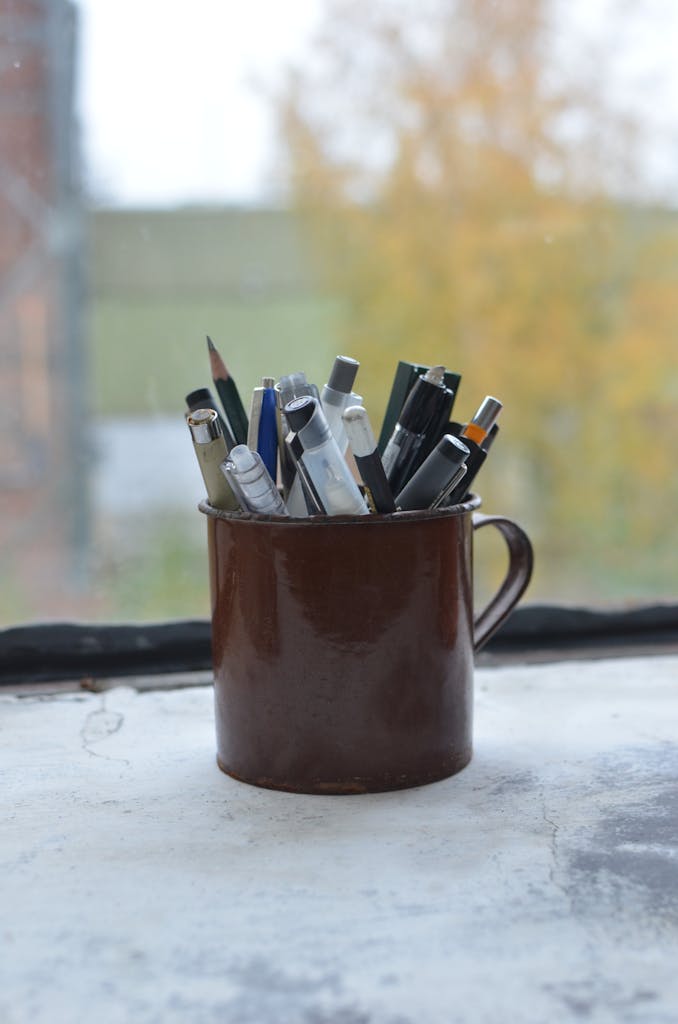 Cup with various stationery consisting of pencils and pens placed on windowsill near window in art workshop