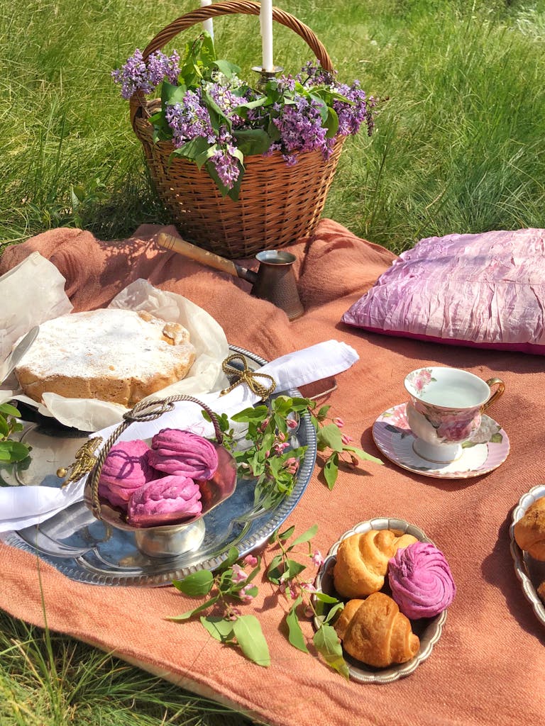 From above of wicker basket with lilac flowers placed on plaid with assorted sweet desserts and porcelain cup during picnic on green grass