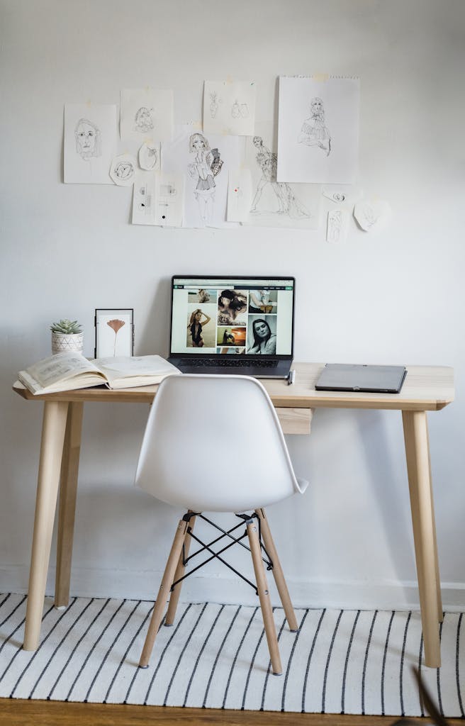 Opened book and laptop with photos on screen with graphic tablet and potted plant placed on wooden table near white wall with hanging sketches in daylight