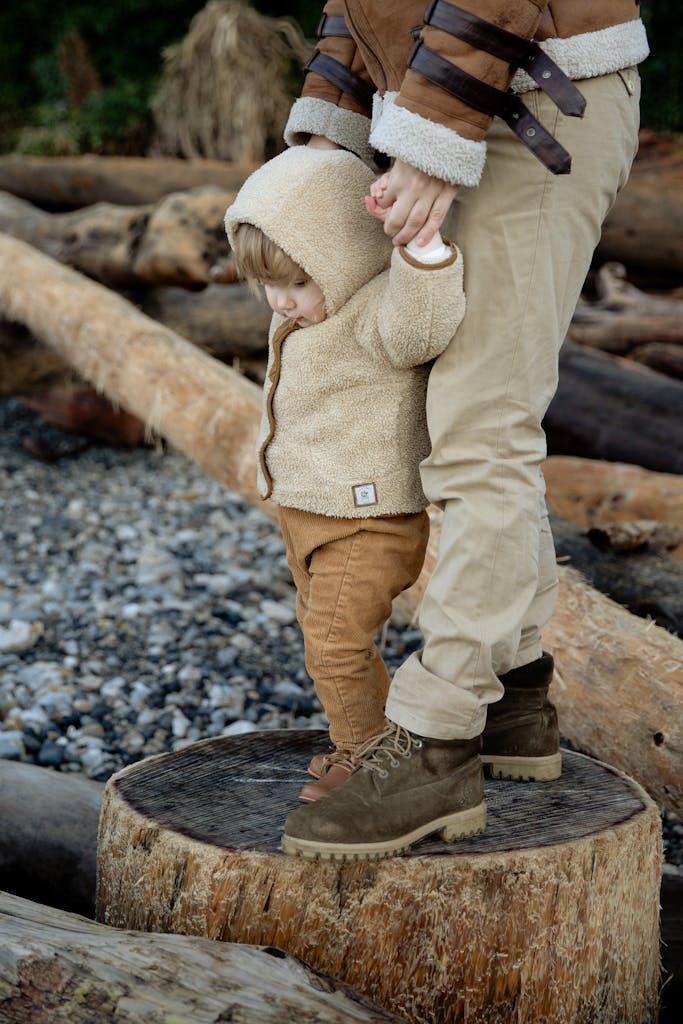 Side view of cute toddler standing with parent on wide log and wearing warm outerwear while spending time together in winter vacation