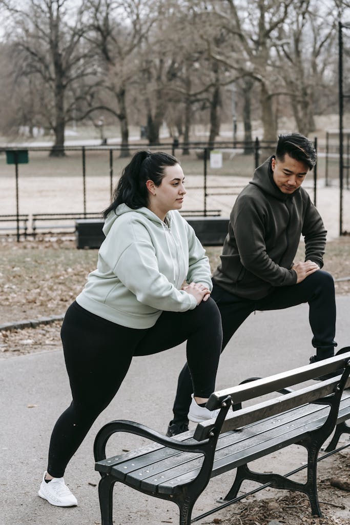 Side view of plus size woman with Asian male personal trainer in sportswear stretching legs on bench during outdoor workout in city park