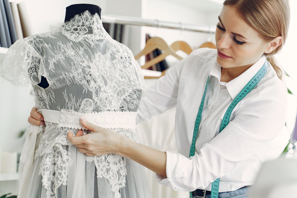 Talented female tailor wearing white shirt and jeans working with mannequin in modern atelier or wedding boutique while attaching belt on lace dress