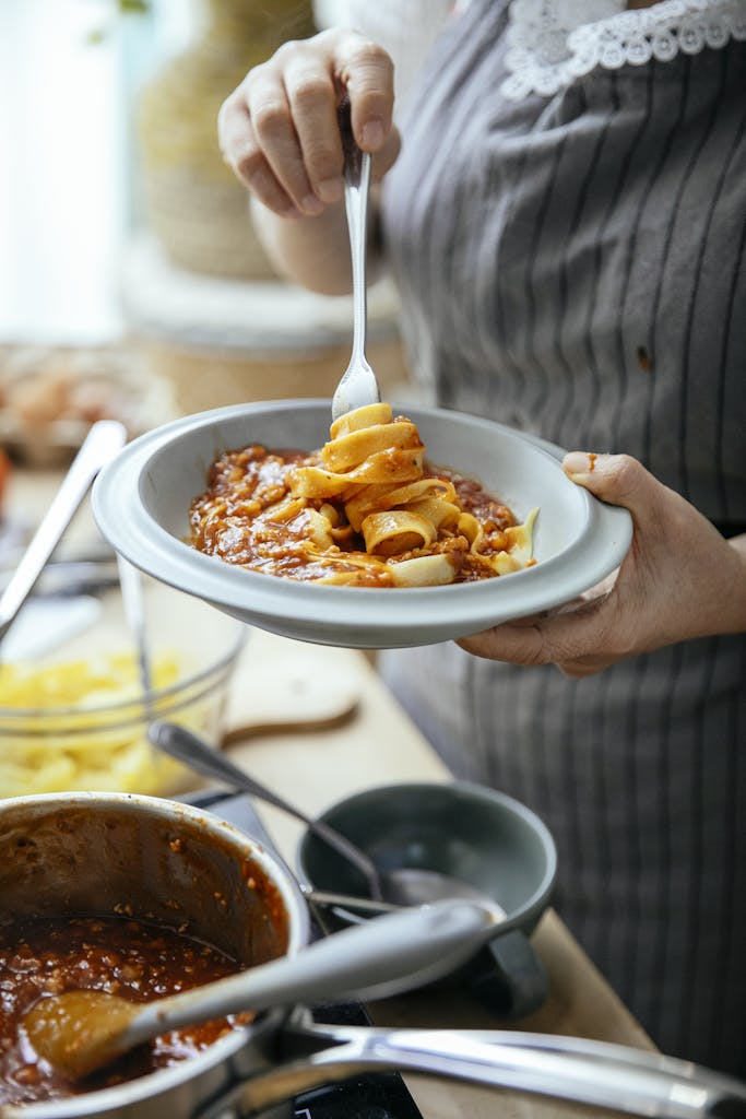 Unrecognizable female cook with plate of pasta wrapped around fork standing at table with pan of tasty chili against blurred background